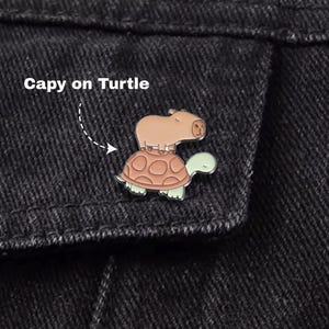 May include: A colorful enamel pin featuring a capybara riding on a turtle. The capybara is brown, and the turtle has a brown shell and green head and feet. The pin is set against a black denim jacket. Text on the image reads "Capy on Turtle."