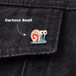 May include: A Cartoon Snail enamel pin with a pink and blue shell, large googly eyes, and a blue wave base. The pin is attached to a black denim jacket. The text "Cartoon Snail" is above the pin.