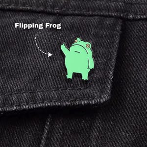 May include: A light green enamel pin in the shape of a frog, giving the middle finger. The pin is attached to a black denim jacket. The text "Flipping Frog" is above the pin.