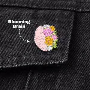 Op de afbeelding: Een speld met een roze hersenontwerp met kleurrijke bloemen die uit één kant bloeien. De tekst "Blooming Brain" staat boven de speld. De speld is geplaatst op een zwarte spijkerjas.