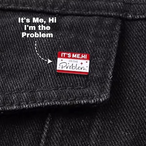 Op de afbeelding: Rode en witte emaille pin met de tekst "It's Me, Hi I'm the Problem" op een zwarte spijkerjas. De pin is rechthoekig met een witte achtergrond en zwarte tekst. De tekst staat ook boven de pin.
