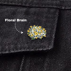 Pode incluir: Close-up de uma jaqueta jeans preta com um broche "Floral Brain". O broche, em forma de cérebro, apresenta flores amarelas e folhas verdes. O texto "Floral Brain" está acima.