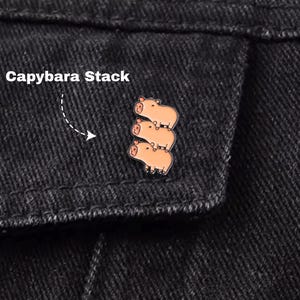 May include: A pin featuring a stack of three cartoon capybaras in shades of brown and tan. The pin is attached to a black denim jacket. The text "Capybara Stack" is visible in the upper left corner.