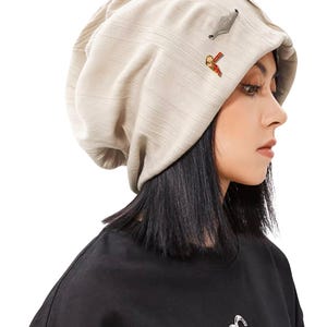 May include: A beige beanie hat with a textured fabric. The hat has two small decorative pins. The person in the image is wearing a black top.