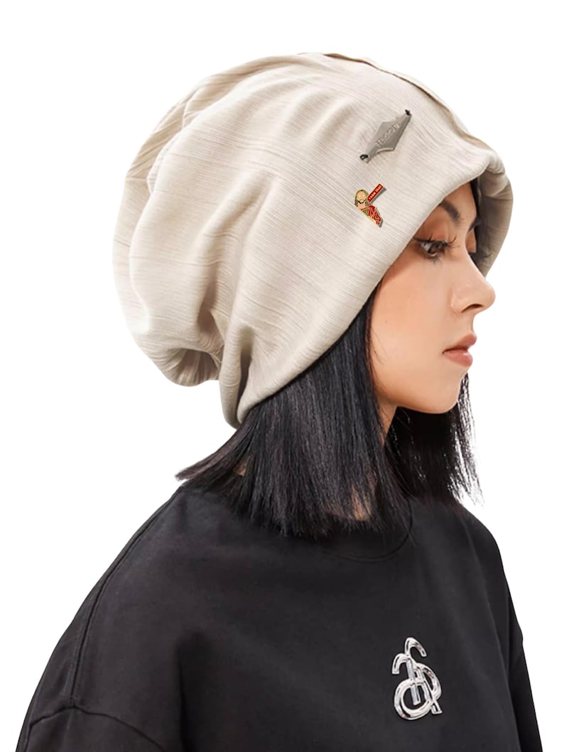 May include: A beige beanie hat with a textured fabric. The hat has two small decorative pins. The person in the image is wearing a black top.