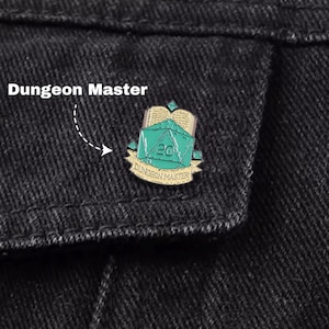 May include: A Dungeon Master enamel pin featuring a green twenty-sided die, an open book, and a banner that reads "Dungeon Master." The pin is attached to a black denim jacket.