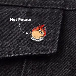 Może przedstawiać: Metalowa przypinka przedstawiająca kreskówkowego ziemniaka w okularach przeciwsłonecznych i płomieniach, z napisem "Hot Potato". Przypinka jest przymocowana do czarnej kurtki dżinsowej.