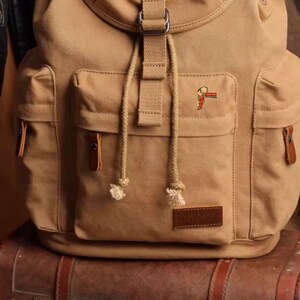 May include: A tan canvas backpack with multiple pockets, a flap closure, and adjustable straps. The backpack has a small pin on the front pocket. The backpack is sitting on top of a brown leather suitcase.