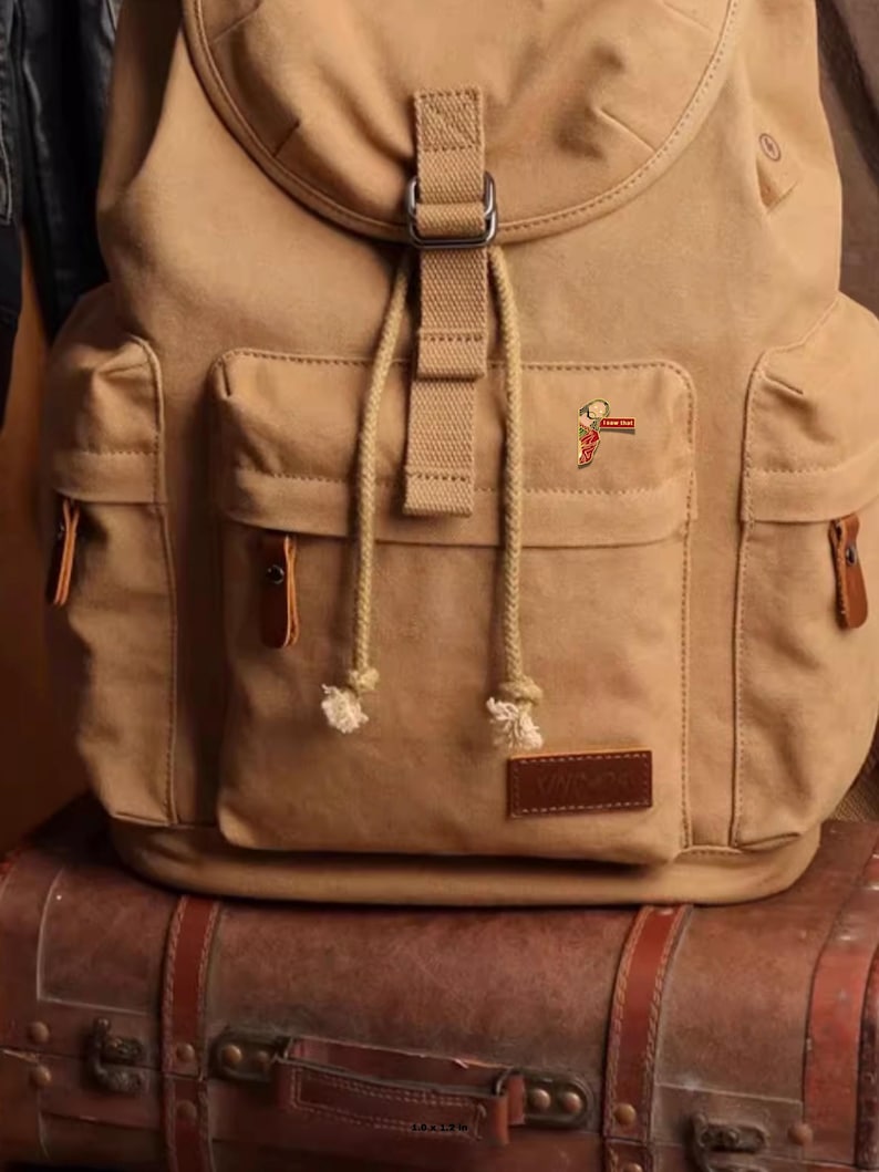 May include: A tan canvas backpack with multiple pockets, a flap closure, and adjustable straps. The backpack has a small pin on the front pocket. The backpack is sitting on top of a brown leather suitcase.