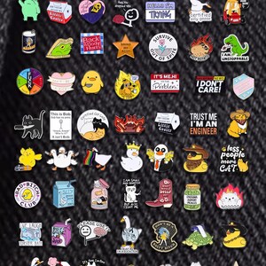 May include: A selection of enamel pins with diverse designs, such as cartoon characters and humorous phrases. The pins display text like "Hello I'm Trying", "I Don't Care!", and "Bad Bitch Club". The pins are in various colors.