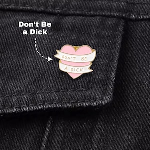 Könnte beinhalten: Ein herzförmiger Emaille-Pin in Rosa mit einem weißen Banner mit der Aufschrift "Don't Be a Dick". Der Pin ist auf einer dunklen Jeansjacke angebracht.