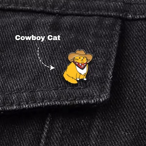 May include: A yellow enamel pin of a cat wearing a brown cowboy hat and a red bandana. The pin is attached to a black denim jacket. The text "Cowboy Cat" is above the pin.