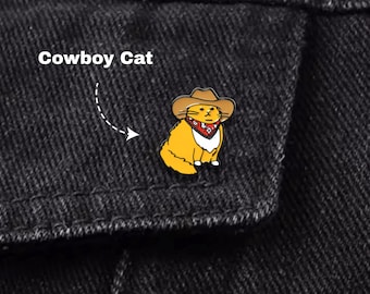 May include: A yellow enamel pin of a cat wearing a brown cowboy hat and a red bandana. The pin is attached to a black denim jacket. The text "Cowboy Cat" is above the pin.