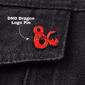 Könnte beinhalten: Ein roter DND Dragon Logo Pin ist an einer schwarzen Jeansjacke befestigt. Der Pin zeigt ein stilisiertes Drachendesign und das kaufmännische Und-Zeichen. Der Text "DND Dragon Logo Pin" wird über dem Pin angezeigt.