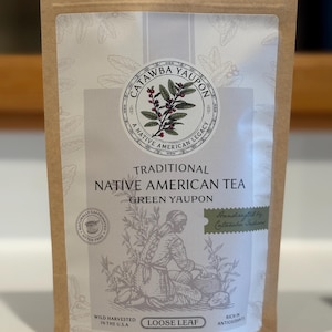 Può includere: Un sacchetto di carta marrone di tè sfuso Catawba Yaupon. L'etichetta presenta un emblema circolare con testo e un'illustrazione botanica. Il testo sull'etichetta recita "Traditional Native American Tea" e "Green Yaupon". La busta contiene 57 g di tè.
