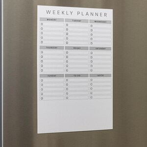 Puede incluir: Un planificador semanal magnético blanco con un diseño sencillo. El planificador es rectangular y presenta secciones para cada día de la semana, una lista de tareas y notas. El texto "WEEKLY PLANNER" está en la parte superior.