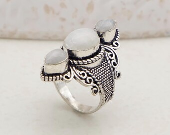 May include: A silver ring with three oval, white gemstones. The ring has an ornate design with a textured band and decorative swirls. The gemstones are set in a bezel setting, and the ring is likely made of sterling silver.
