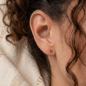Puede incluir: Primer plano de una oreja con un peque&ntilde;o pendiente de bot&oacute;n dorado con la letra "K". El pendiente tiene un dise&ntilde;o sencillo y minimalista. La persona tiene el pelo casta&ntilde;o rizado y lleva una camiseta color crema.