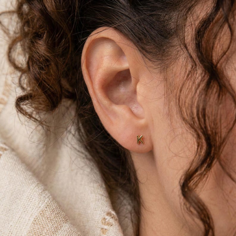 Puede incluir: Primer plano de una oreja con un peque&ntilde;o pendiente de bot&oacute;n dorado con la letra "K". El pendiente tiene un dise&ntilde;o sencillo y minimalista. La persona tiene el pelo casta&ntilde;o rizado y lleva una camiseta color crema.