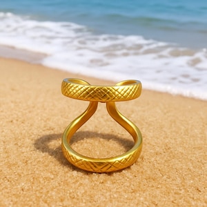 Puede incluir: Un anillo dorado con un diseño único de doble banda, colocado en una playa arenosa. El anillo presenta una superficie texturizada con un patrón de diamantes. El océano y el cielo son visibles en el fondo, creando un tema de playa.