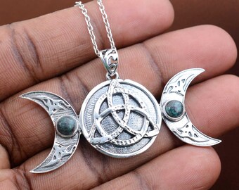 May include: A silver-colored pendant necklace featuring a central circular design with a Celtic knot, flanked by crescent moons. Each moon and the central circle has a small, round, teal-colored stone. The necklace is held by a hand.