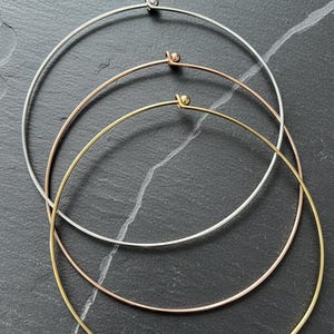Può includere: Tre collane in metallo argentato, oro rosa e oro. Ogni collana è un semplice cerchio di filo sottile con una piccola chiusura. Le collane sono disposte su una superficie scura e strutturata.