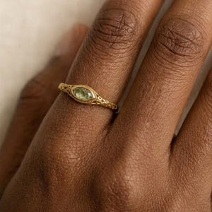 Puede incluir: Un anillo dorado con una piedra preciosa ovalada de color verde claro. El anillo presenta un intrincado diseño calado alrededor de la piedra preciosa y a lo largo de la banda. El anillo está en un dedo.