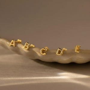 May include: Gold-coloured letter stud earrings, spelling out the letters A, B, C, D, and E. The earrings are displayed on a light-coloured, textured surface. The letters are in a serif font.