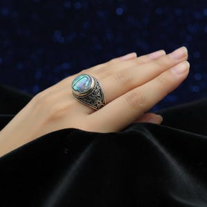 May include: A silver ring with an oval, iridescent abalone shell stone. The ring has an ornate, filigree design around the stone and on the band. The ring is displayed on a hand against a dark blue background.