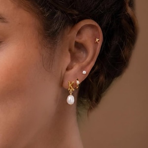 Puede incluir: Primer plano de una oreja adornada con pendientes dorados. Los pendientes incluyen un peque&ntilde;o pendiente de bot&oacute;n, un aro con una peque&ntilde;a gema, un dije con la letra "K" y una perla colgante. El fondo es borroso y neutro.