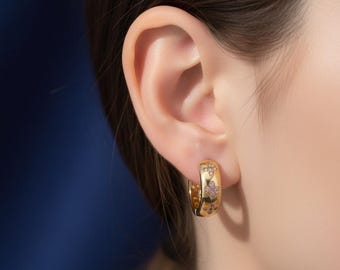May include: Close-up of a gold hoop earring with a floral design and small pink gemstones. The earring is worn on an ear, with the focus on the jewelry's details and the ear's shape. The earring has a polished finish.