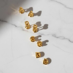 Puede incluir: Pendientes de bot&oacute;n dorados con forma de letras, dispuestos sobre una superficie de m&aacute;rmol blanco. Se muestran las letras A, B, C y D, cada una en pares. Los pendientes tienen un acabado brillante y proyectan sombras.