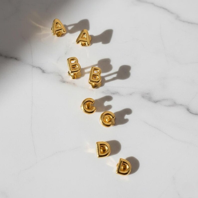 Puede incluir: Pendientes de bot&oacute;n dorados con forma de letras, dispuestos sobre una superficie de m&aacute;rmol blanco. Se muestran las letras A, B, C y D, cada una en pares. Los pendientes tienen un acabado brillante y proyectan sombras.