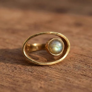 Puede incluir: Un anillo dorado con un diseño ovalado y una piedra preciosa redonda e iridiscente. La piedra preciosa está engastada en un bisel y es el punto focal del anillo. El anillo está sobre una superficie de madera.