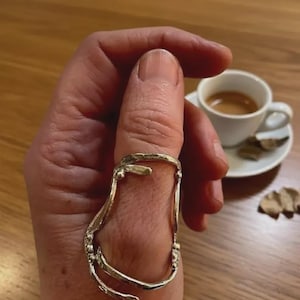 Puede incluir: Un anillo plateado con un diseño alargado inusual, usado en un pulgar. El anillo tiene una apariencia texturizada y orgánica. Una pequeña taza blanca de café está en el fondo sobre un platillo.