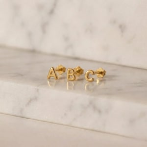 Puede incluir: Tres pendientes de bot&oacute;n dorados que deletrean las letras "A", "B" y "C". Los pendientes son peque&ntilde;os y tienen un dise&ntilde;o simple y elegante. Se colocan sobre una superficie de m&aacute;rmol blanco y gris.