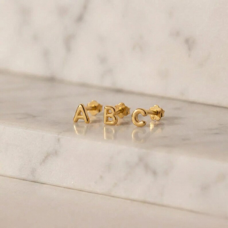 Puede incluir: Tres pendientes de bot&oacute;n dorados que deletrean las letras "A", "B" y "C". Los pendientes son peque&ntilde;os y tienen un dise&ntilde;o simple y elegante. Se colocan sobre una superficie de m&aacute;rmol blanco y gris.