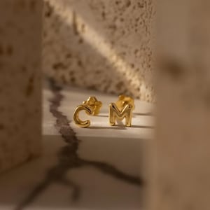 Puede incluir: Un par de pendientes dorados con forma de letras "C" y "M". Los pendientes se muestran sobre una superficie blanca con un fondo borroso.
