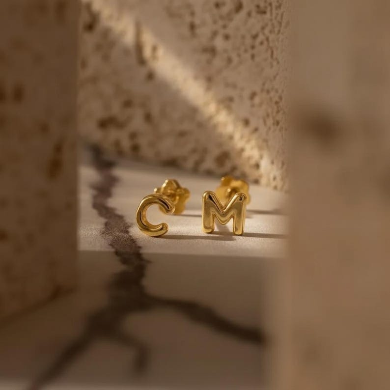 Puede incluir: Un par de pendientes dorados con forma de letras "C" y "M". Los pendientes se muestran sobre una superficie blanca con un fondo borroso.