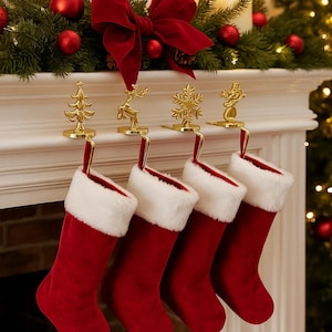 Può includere: Quattro calze natalizie in velluto rosso con polsini in finta pelliccia bianca sono appese a un camino. I porta calze dorati presentano un albero di Natale, una renna, un fiocco di neve e un pupazzo di neve. Un fiocco rosso e ornamenti decorano il camino.