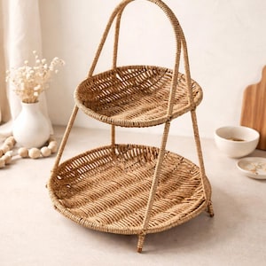 Può includere: Un vassoio da portata a due livelli in vimini naturale con manico. Il vassoio è di forma rotonda. Il manico si incurva sopra il livello superiore. Il vassoio è sostenuto da gambe sottili.