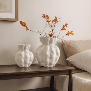 May include: Two textured white vases with a ribbed design sit on a dark wood table. One vase is smaller than the other. Orange-colored branches with leaves are in the larger vase. The scene is set in a bedroom.