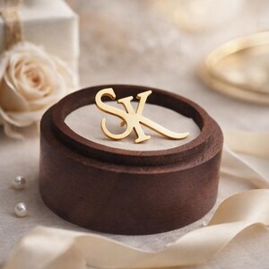 Könnte beinhalten: Goldfarbene Monogramm-Anstecknadel mit den Buchstaben "SK" in einer dunkelbraunen Holzringbox mit cremefarbenem Stoff. Eine weiße Rose, Perlen und ein goldenes Armband sind im Hintergrund, mit einem cremefarbenen Band.