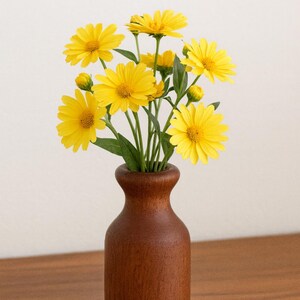 May include: A wooden vase holds a bouquet of bright yellow flowers with green stems and leaves. The vase is a dark brown color and sits on a wooden surface. The flowers have multiple petals and are in full bloom.