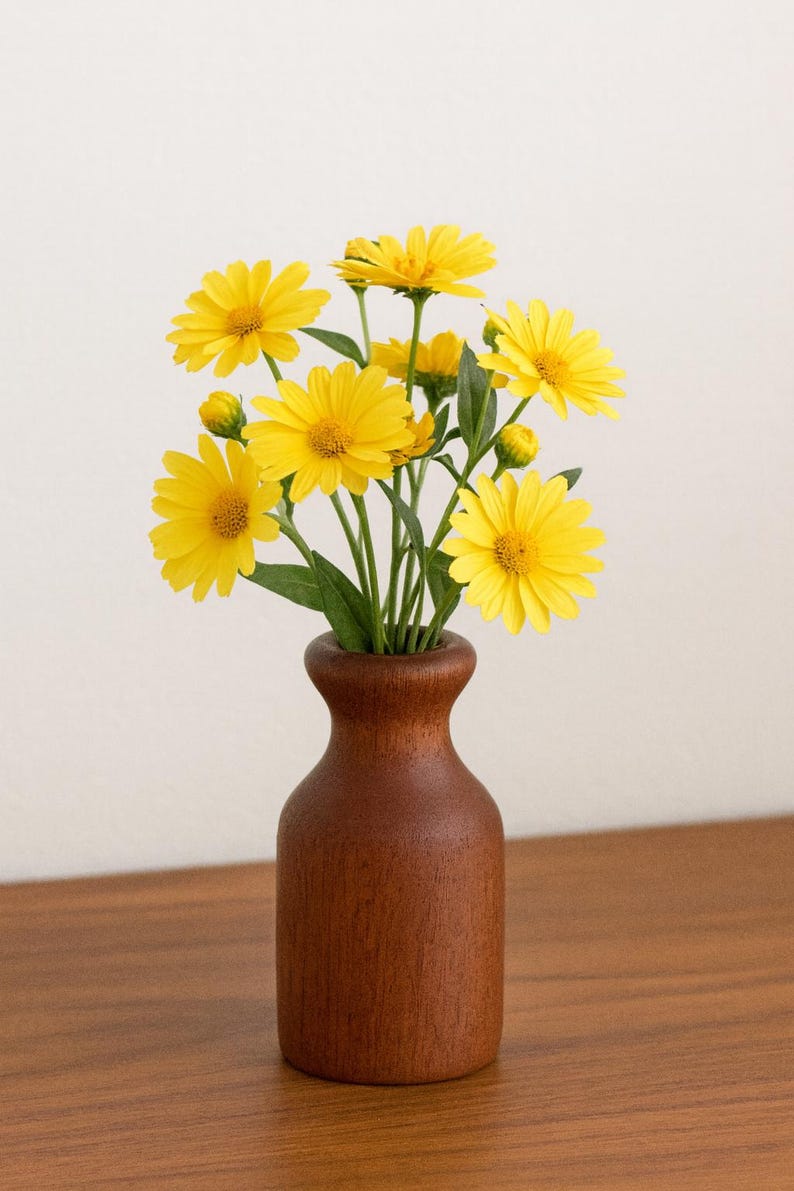 May include: A wooden vase holds a bouquet of bright yellow flowers with green stems and leaves. The vase is a dark brown color and sits on a wooden surface. The flowers have multiple petals and are in full bloom.