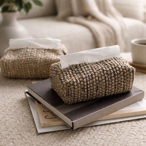 May include: Two woven tissue box covers in a neutral color, one with tissues partially pulled out. The boxes are rectangular and sit on a stack of books. The boxes have a textured, nubby appearance, and the background is a soft, neutral color.