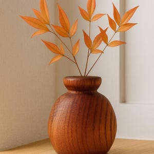 May include: A small, round wooden vase with a narrow neck holds several sprigs of orange leaves. The vase is made of a dark, reddish-brown wood and sits on a light-colored wooden surface. The leaves have a vibrant orange hue.
