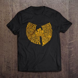 May include: Black t-shirt with a gold-coloured graphic in the shape of a stylized letter W. The graphic is filled with text, including names and phrases. The shirt is displayed on a wooden surface.