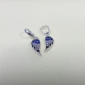 Puede incluir: Dos dijes plateados con forma de alas de ángel, cada uno con una argolla en la parte superior. La superficie de las alas está decorada con un degradado de pequeñas piedras brillantes azules y transparentes, desde el azul oscuro hasta el claro.