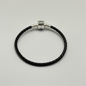 Puede incluir: Pulsera de cuero trenzado negro con cierre plateado. El cierre tiene grabadas las letras "PA". La pulsera es circular y está sobre una superficie blanca. Es una pieza de joyería.